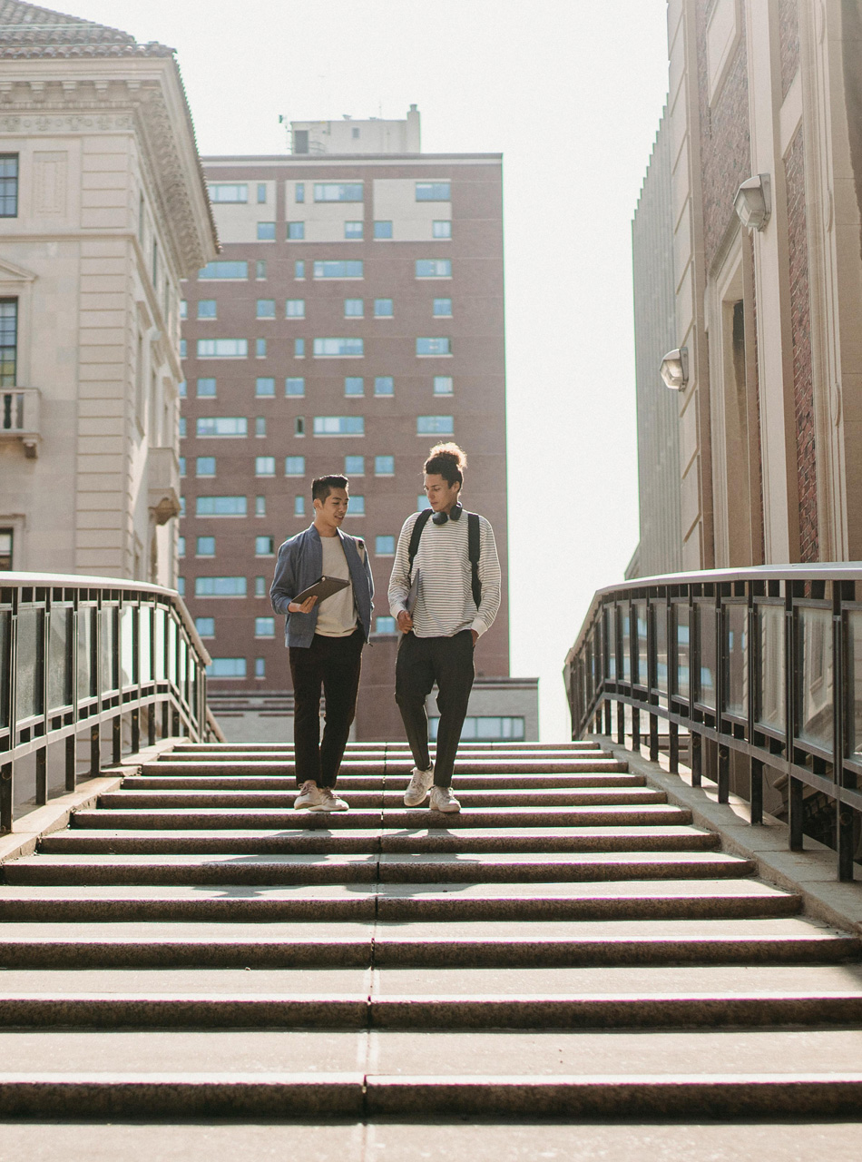 Two college students walking on a campus bridge between historic and modern student housing buildings.